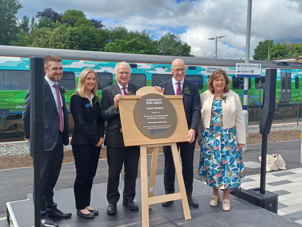 First Minister John Swinney, Transport Secretary Fiona Hyslop and project partners pose for a photo with the new plaque at Leven Station. A specially wrapped train is in the background